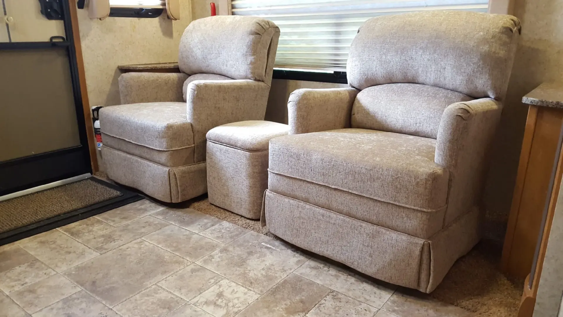 Two beige upholstered recliners with a matching ottoman inside a camper, positioned beside a window.